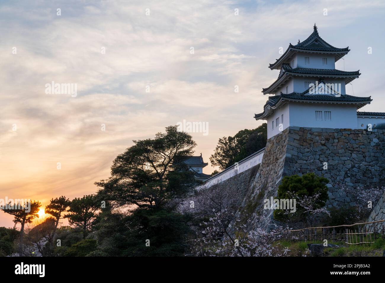 Sunset at Akashi castle in Japan. View along the two Edo Period yagura, turrets, separated by a long stone wall, ishigaki with dobei white plaster wall on top. The sun setting in some trees in a cloudy blue sky. Stock Photo