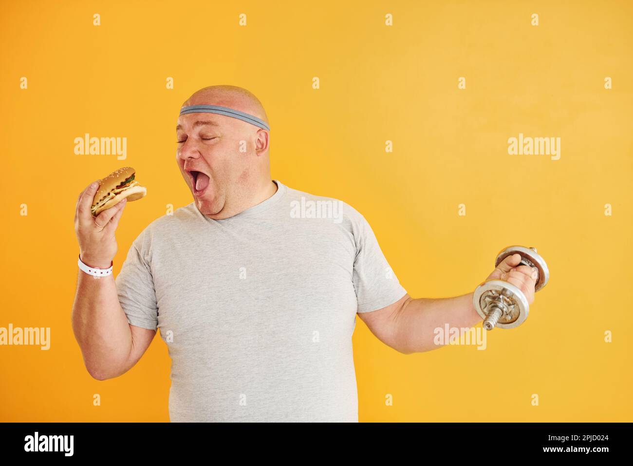 Holds hamburger and dumbbell. Funny overweight man in sportive head tie