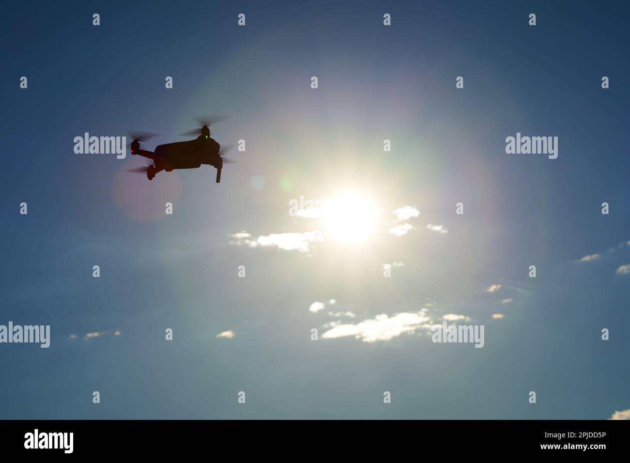 Dark drone from the side as silhouette. Yellow sun in front of dark blue sky with white clouds. Up view. Stock Photo