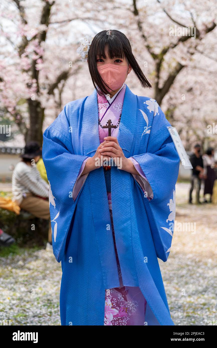 Young Japanese women wearing the costume of a 16th Century Christian holding a wood cross in both hands and wearing a blue cloak and face mask. Standing under sunlit cherry blossoms. Part of the samurai parade held in the springtime cherry blossom festival at Hon-Tatsuno in Japan. Stock Photo