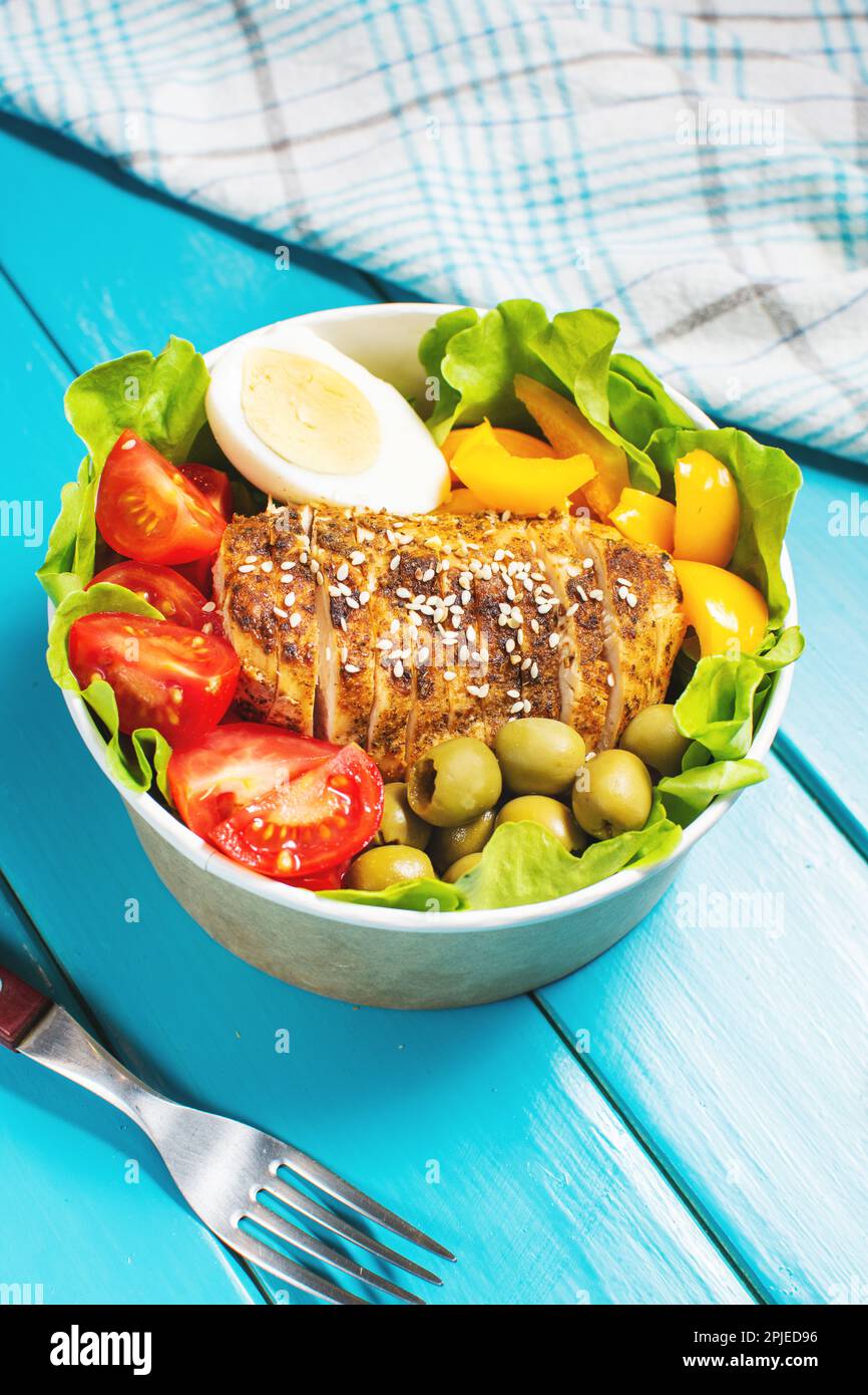 Buddha bowl salad with grilled chicken breast slices, cherry tomatoes, eggs, pepper, olive, lettuce and seeds on blue wooden background. Healthy food, Stock Photo