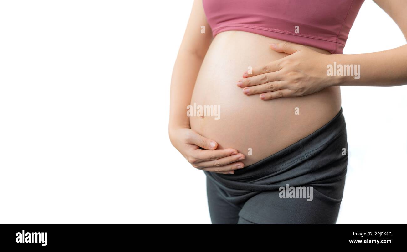 Close up of the happy pregnant woman in fitness clothes standing happily holding her exposed belly against white background, room for copy space Stock Photo