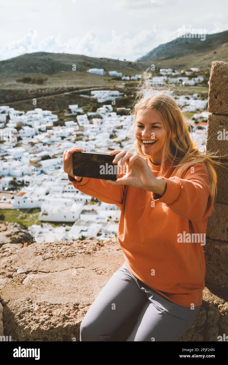 Travel blogger woman taking selfie photo by smartphone camera influencer girl in Greece sightseeing Lindos town aerial view Rhodes island summer vacat Stock Photo