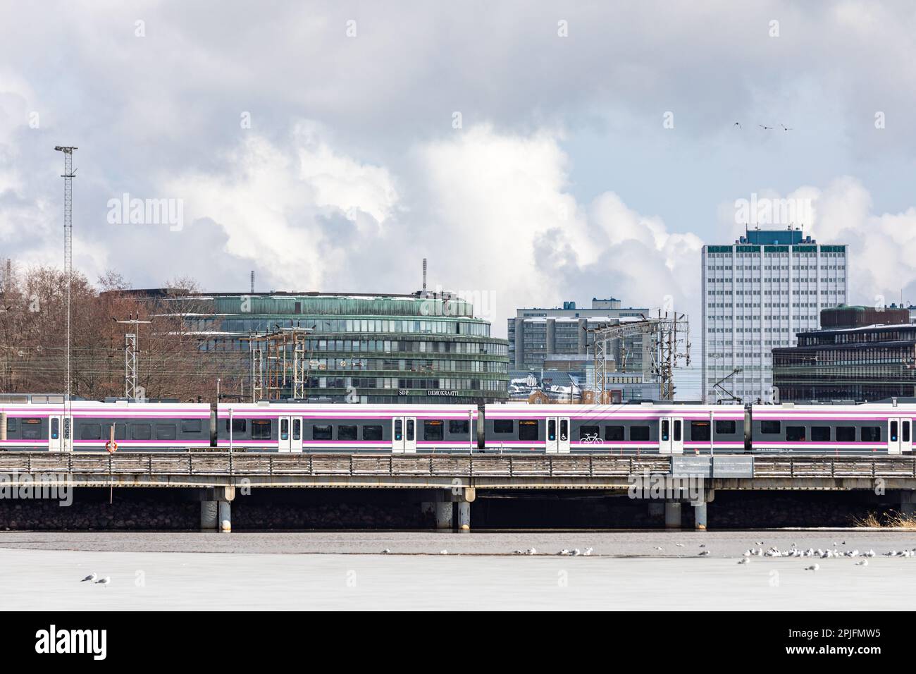 Commuter train passing a railway bridge with Hakaniemi and Merihaka buildings in the background on a cloudy early spring day in Helsinki, Finland Stock Photo