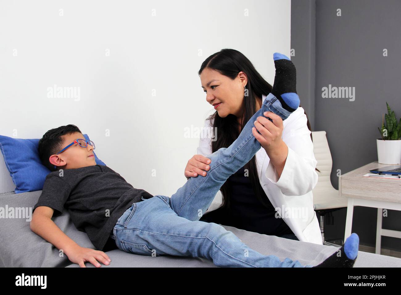 Latina pediatrician woman doctor attends a 7-year-old boy in her office checks his leg, knee and foot for falls and diseases such as rickets, blount Stock Photo