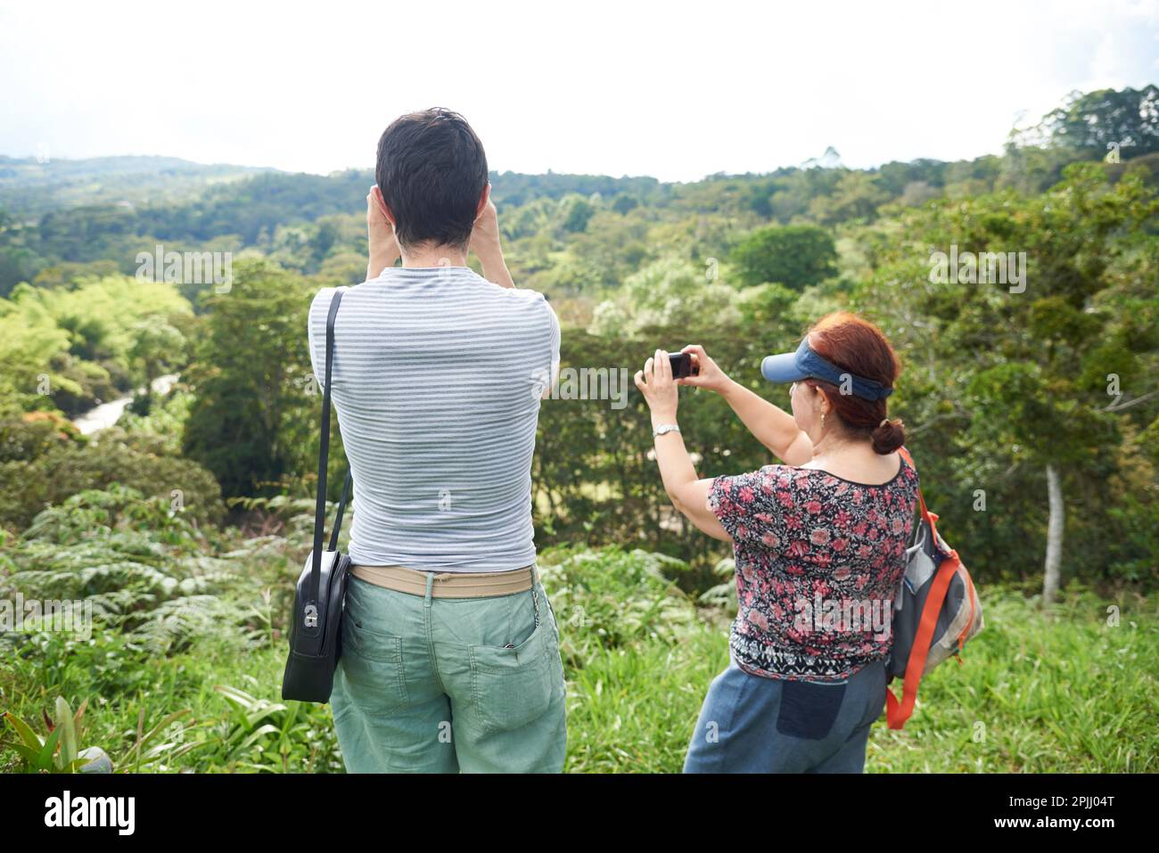 Mother with her adult son enjoying a trip, he looks at the landscape with binoculars while she takes pictures. Stock Photo