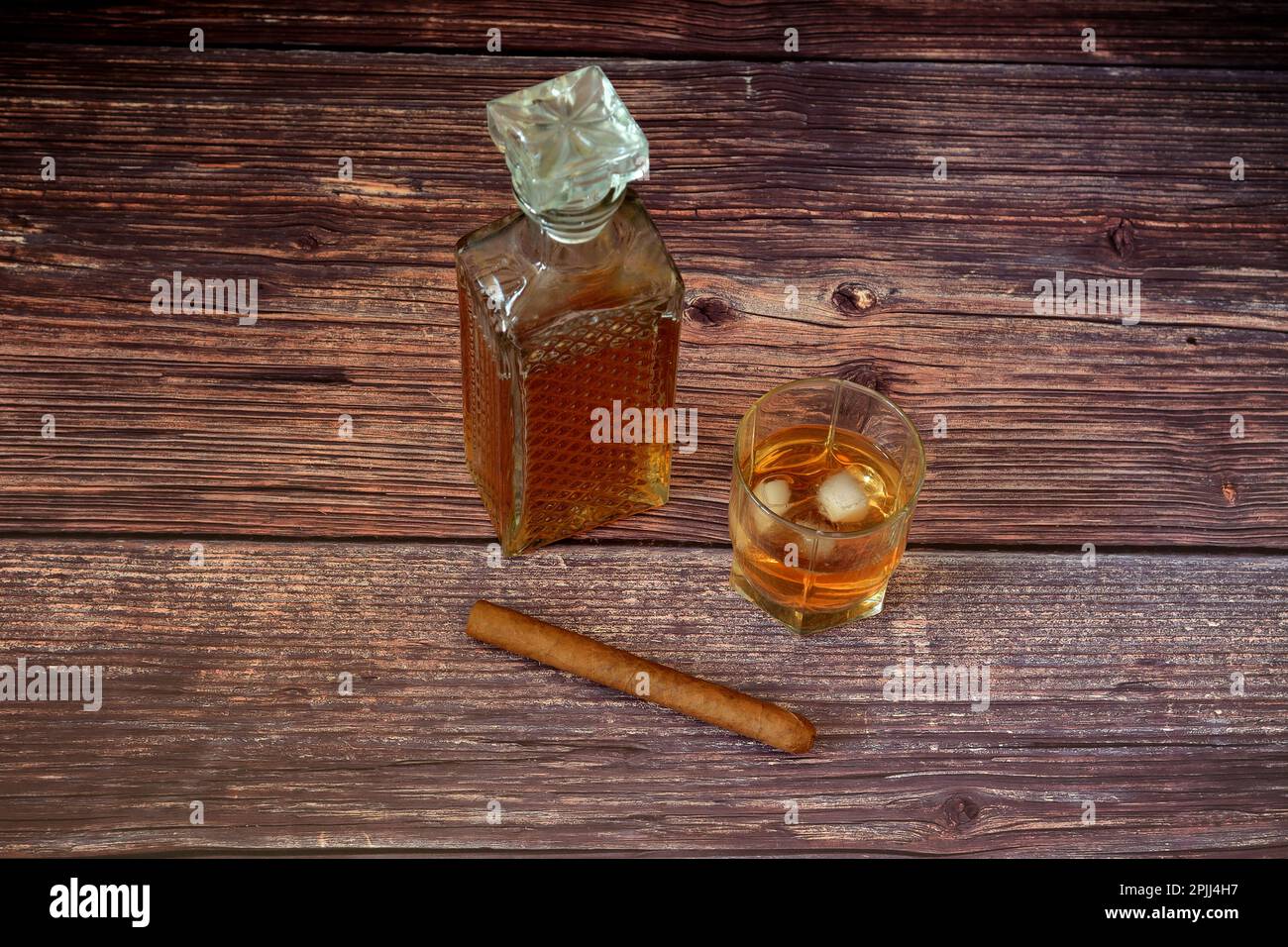 A glass glass of whiskey with ice, a crystal decanter with an alcoholic drink and a Cuban cigar on a wooden table. Close-up. Stock Photo