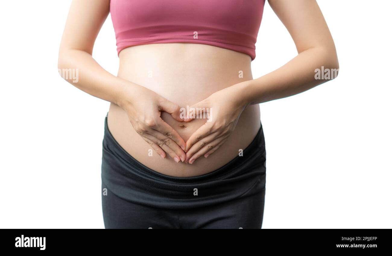 Close up of the happy pregnant woman in fitness clothes standing using her hands to make a heart shape over her exposed belly against white background Stock Photo