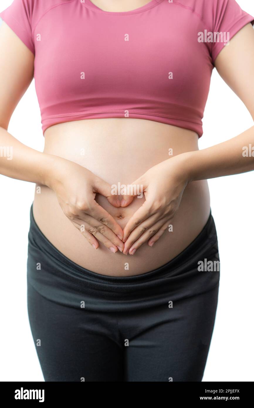 Close up of the happy pregnant woman in fitness clothes standing using her hands to make a heart shape over her exposed belly against white background Stock Photo