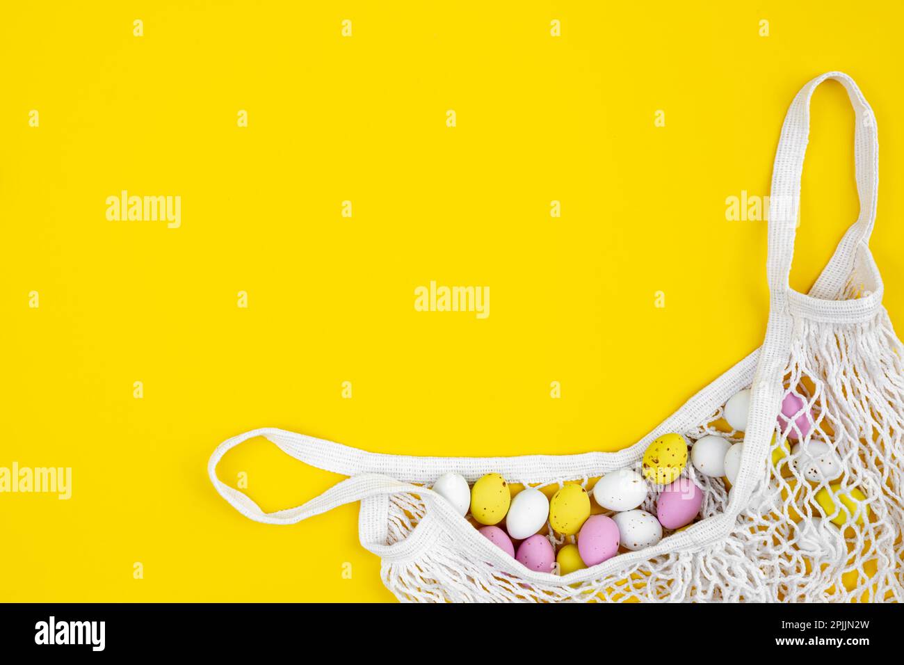 Colored Easter eggs in a mesh bag on a yellow background, flat lay. Stock Photo
