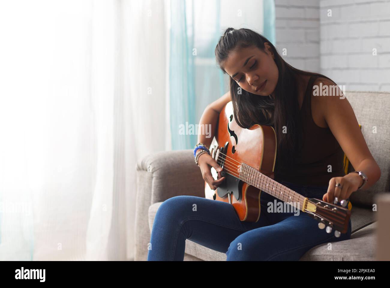 Young woman tuning guitar while sitting on sofa in living room Stock Photo