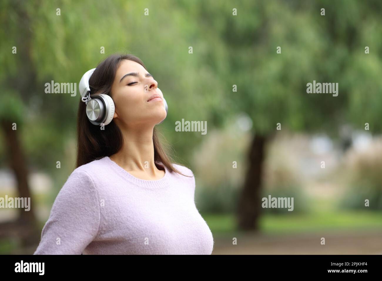 Relaxed woman breathing fresh air with headphone meditating in a park ...