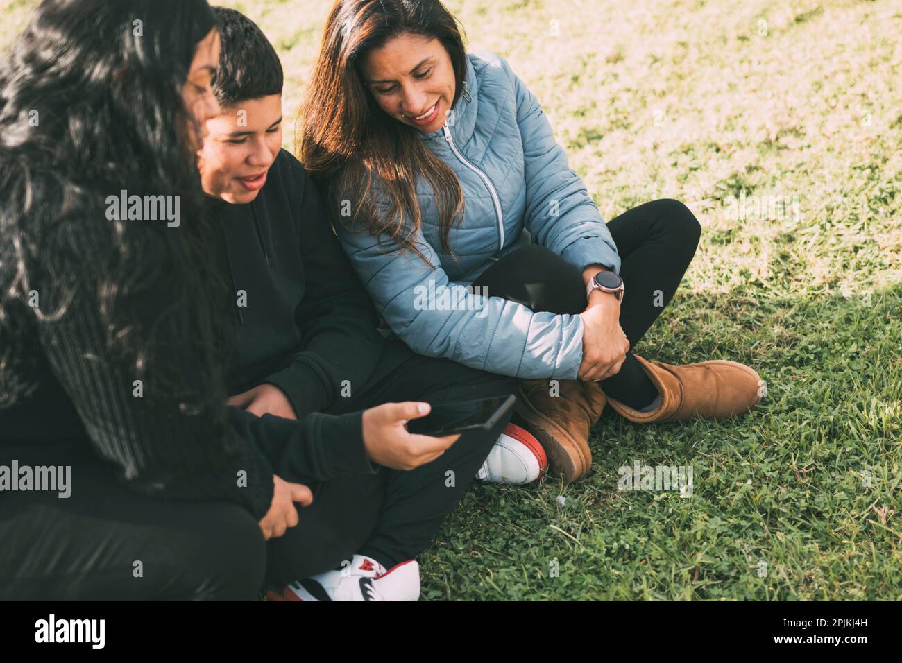 Hispanic family spending quality time together in a local park on a beautiful, sunny day. A teenage boy is sitting on the grass, holding his Stock Photo