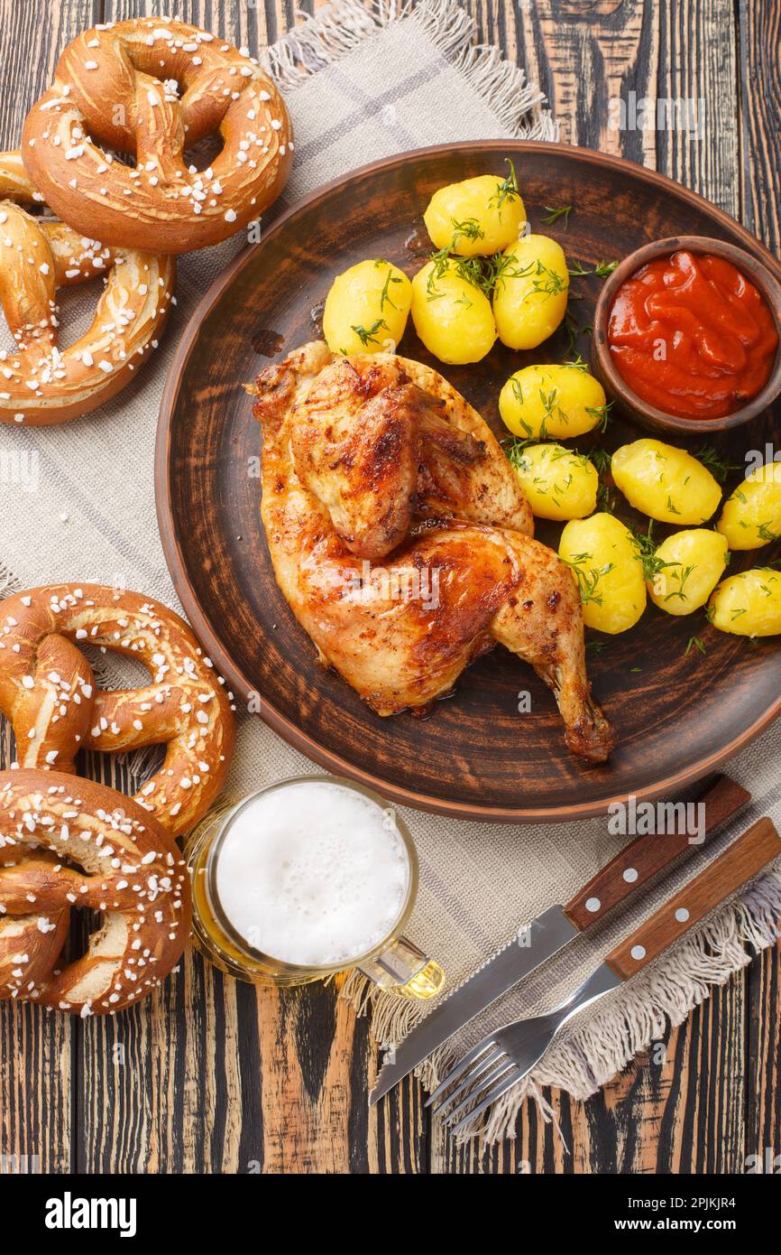 German holiday food half fried chicken with boiled potatoes, sauce, beer and pretzels close-up on a wooden table. Vertical top view from above Stock Photo