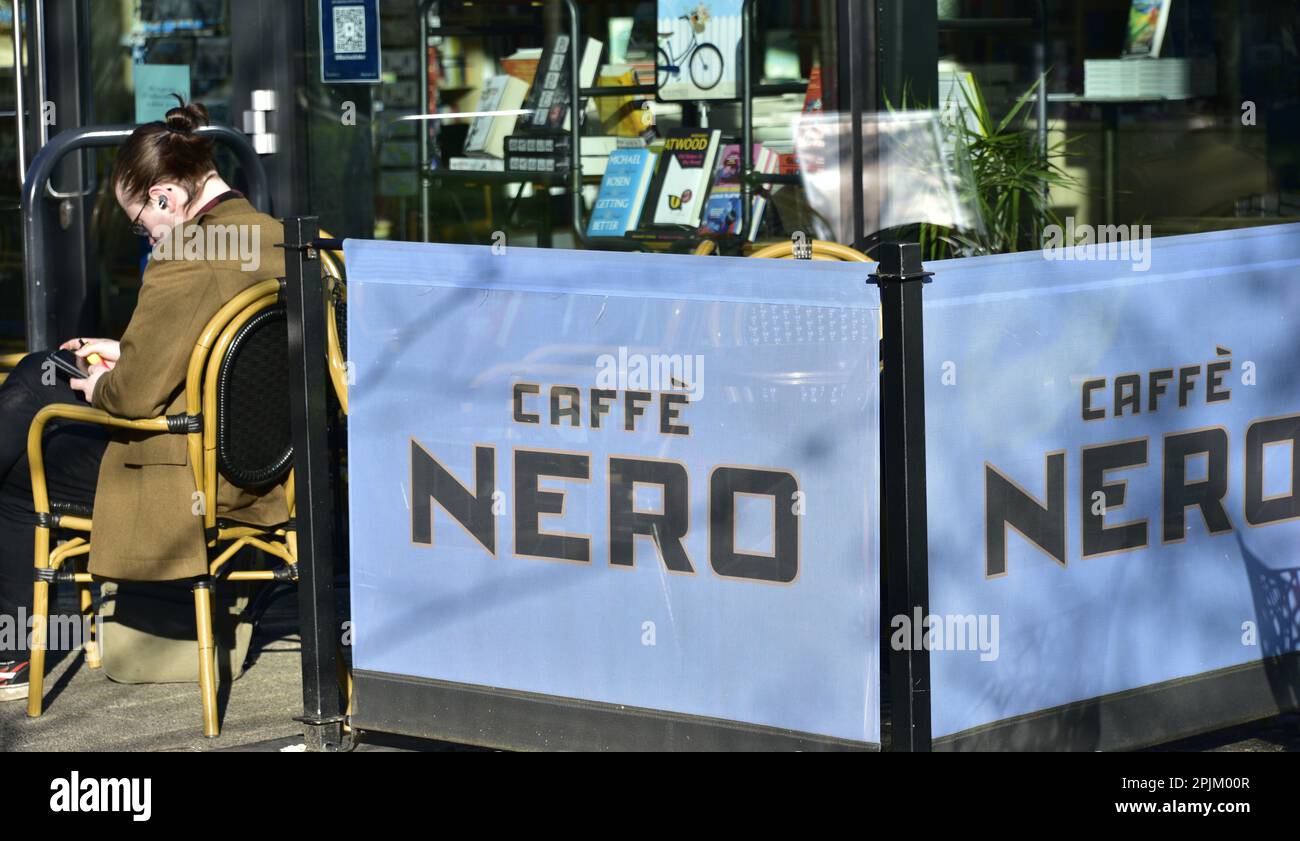 A person sits at a table in the sunshine beside a Caffè Nero sign or notice in Manchester, UK. Stock Photo