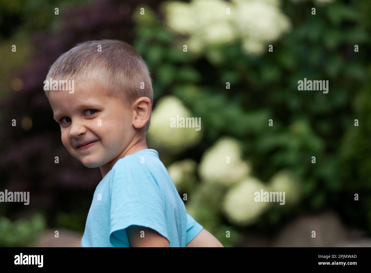 Side portrait of a handsome child, adorable boy in blue t-shirt looking through his shoulder at camera, with summer natur background. Copy ad space Stock Photo