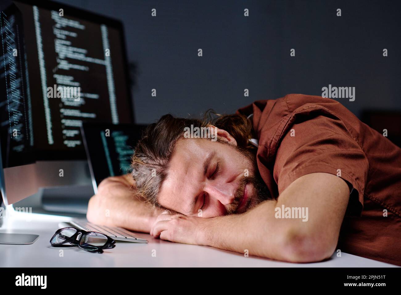 Young exhausted male programmer with his head on desk by computer keyboard sleeping after work over developing new decoding software Stock Photo