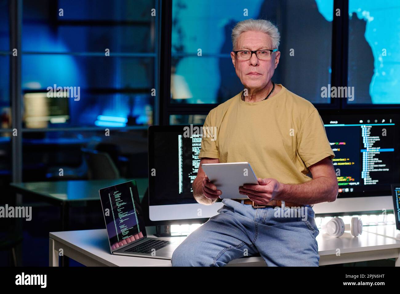 Experienced IT engineer with grey hair using tablet and looking at camera while sitting on desk with computers with coded data on screens Stock Photo