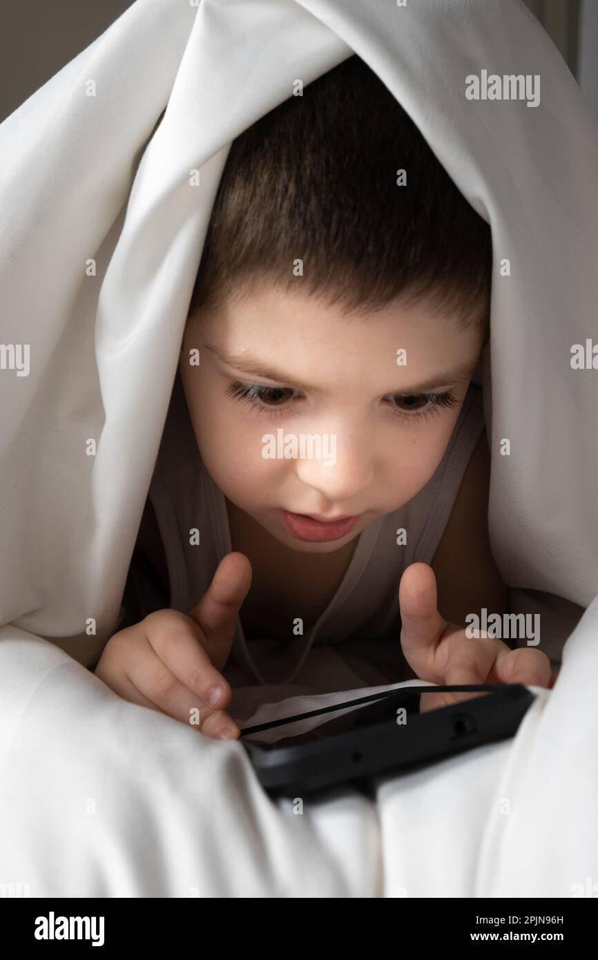 A 5 year old boy plays games on his phone while lying in bed. Gadgets in bed before bedtime in a