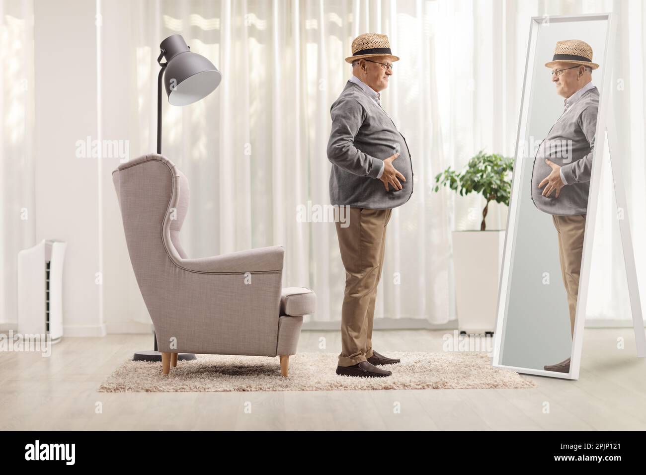 Elderly man with a big belly standing in front of an armchair and looking at a mirror at home Stock Photo