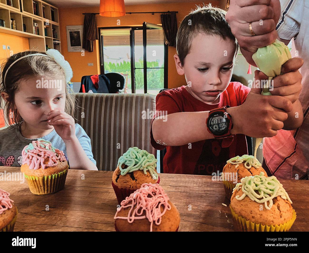 Group of children baking cupcakes, squeezing cream from confectionery bag, preparing ingredients, topping, sprinkles for decorating cookies. Kids cook Stock Photo