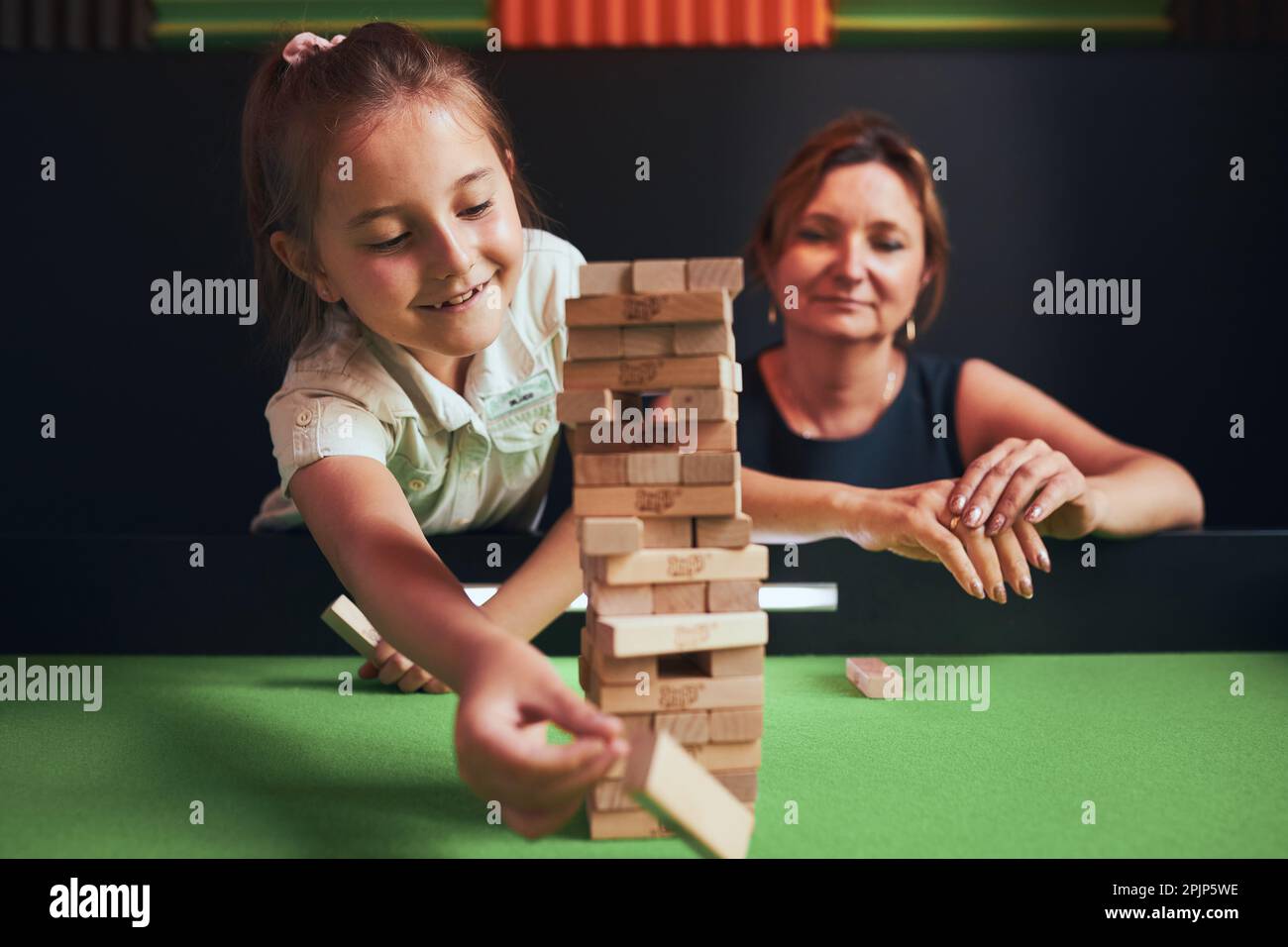 Mother and her daughter playing jenga game together in play room. Girl removing one block from stack and placing it on top of tower. Stacked wooden bl Stock Photo