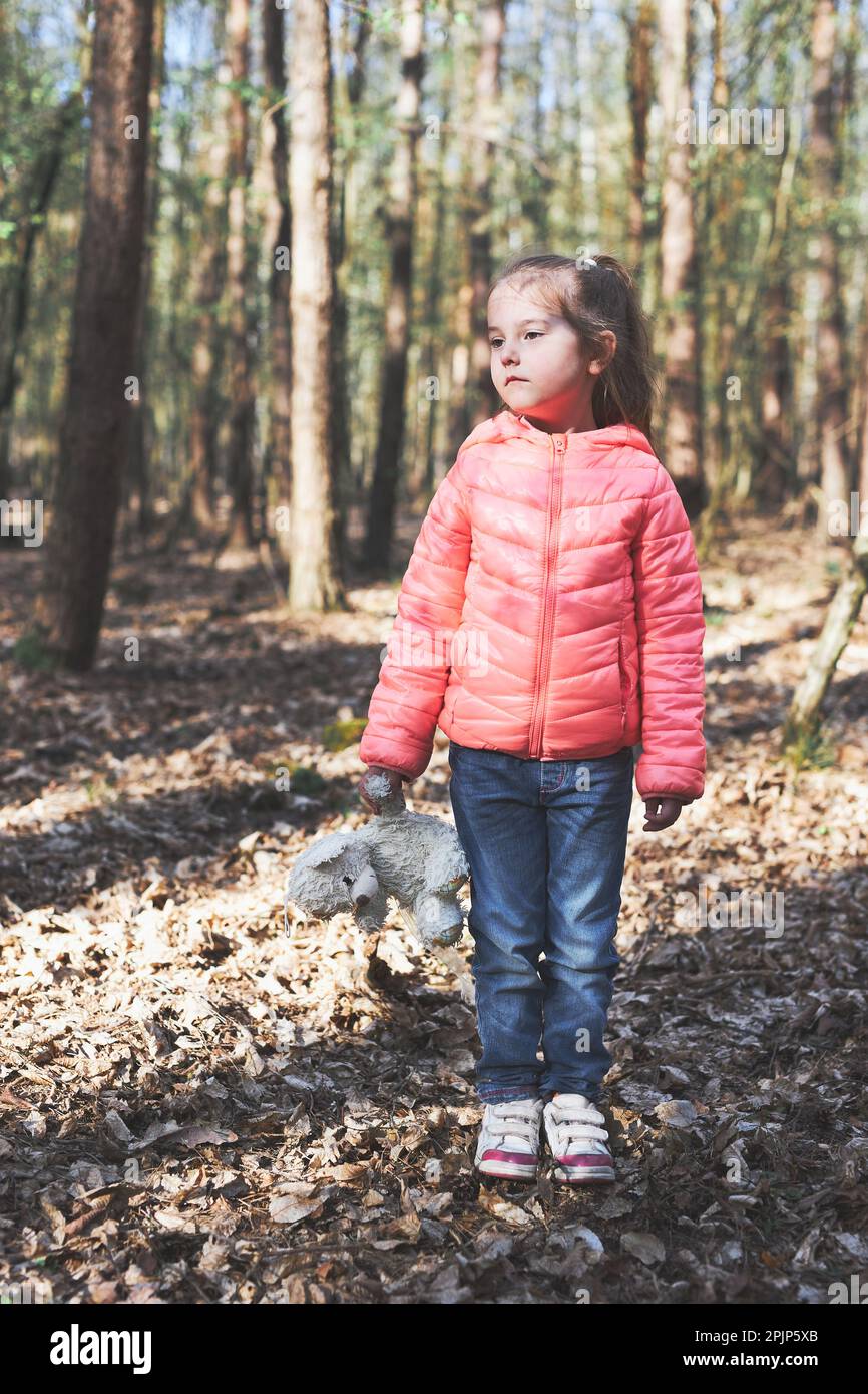 Little girl child standing on stump in a forest during a walk on sunny spring day keeping a toy teddy bear looking away Stock Photo