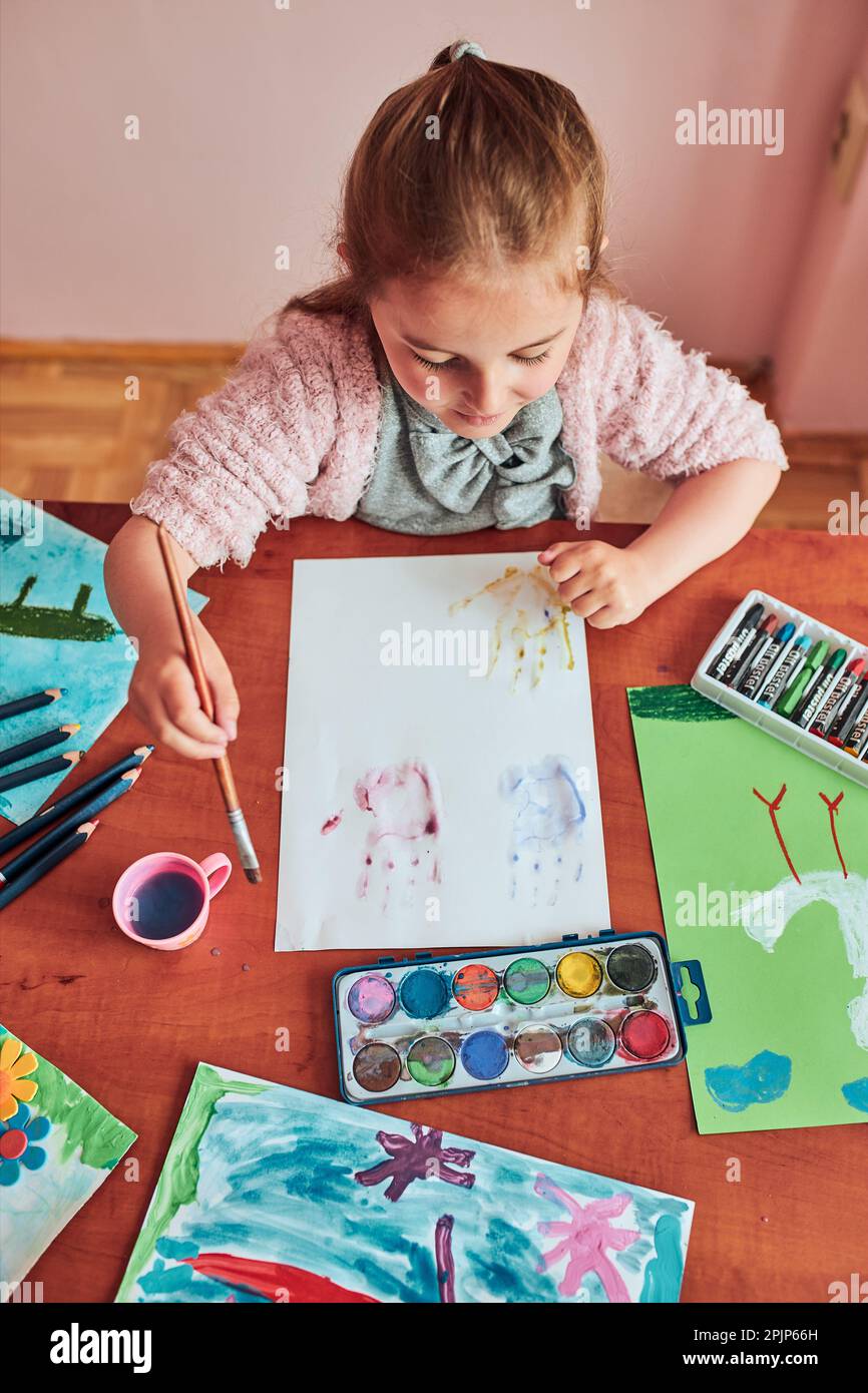 Little girl preschooler painting a picture using colorful paints and crayons. Child having fun making a picture during an art class in the classroom Stock Photo