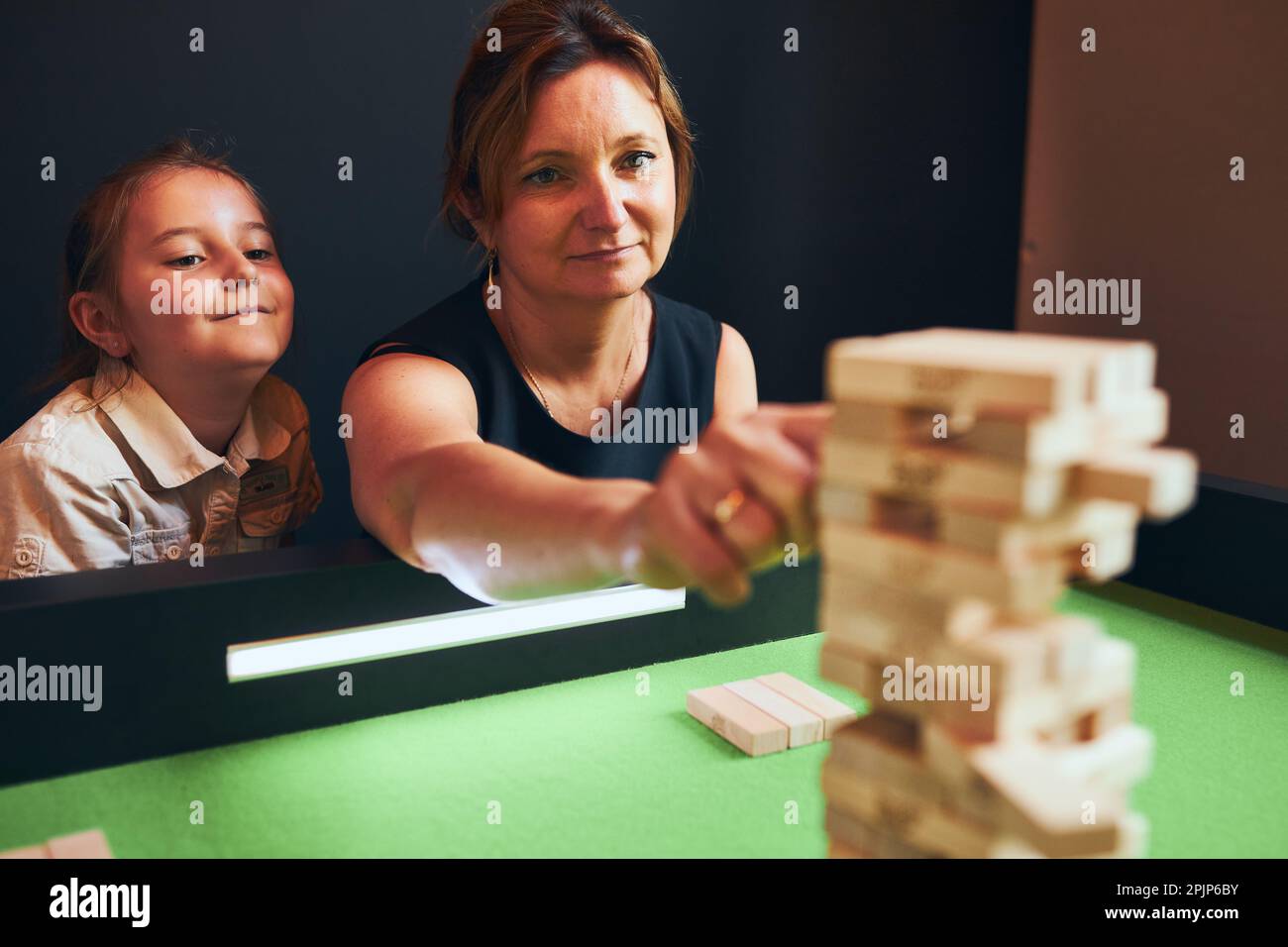 Mother playing jenga game with her daughter in play room. Woman removing block from stack. Unstable tower of wooden blocks. Game of skill and fun. Fam Stock Photo