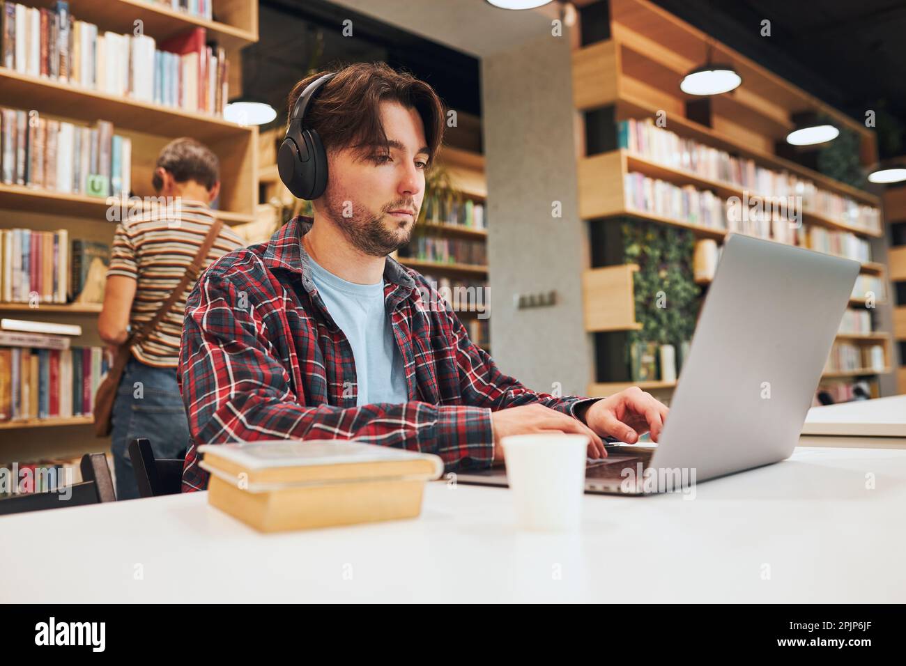 Student learning in university library. Young man preparing for test on laptop. Man listening to online course. Focused student studying for college e Stock Photo