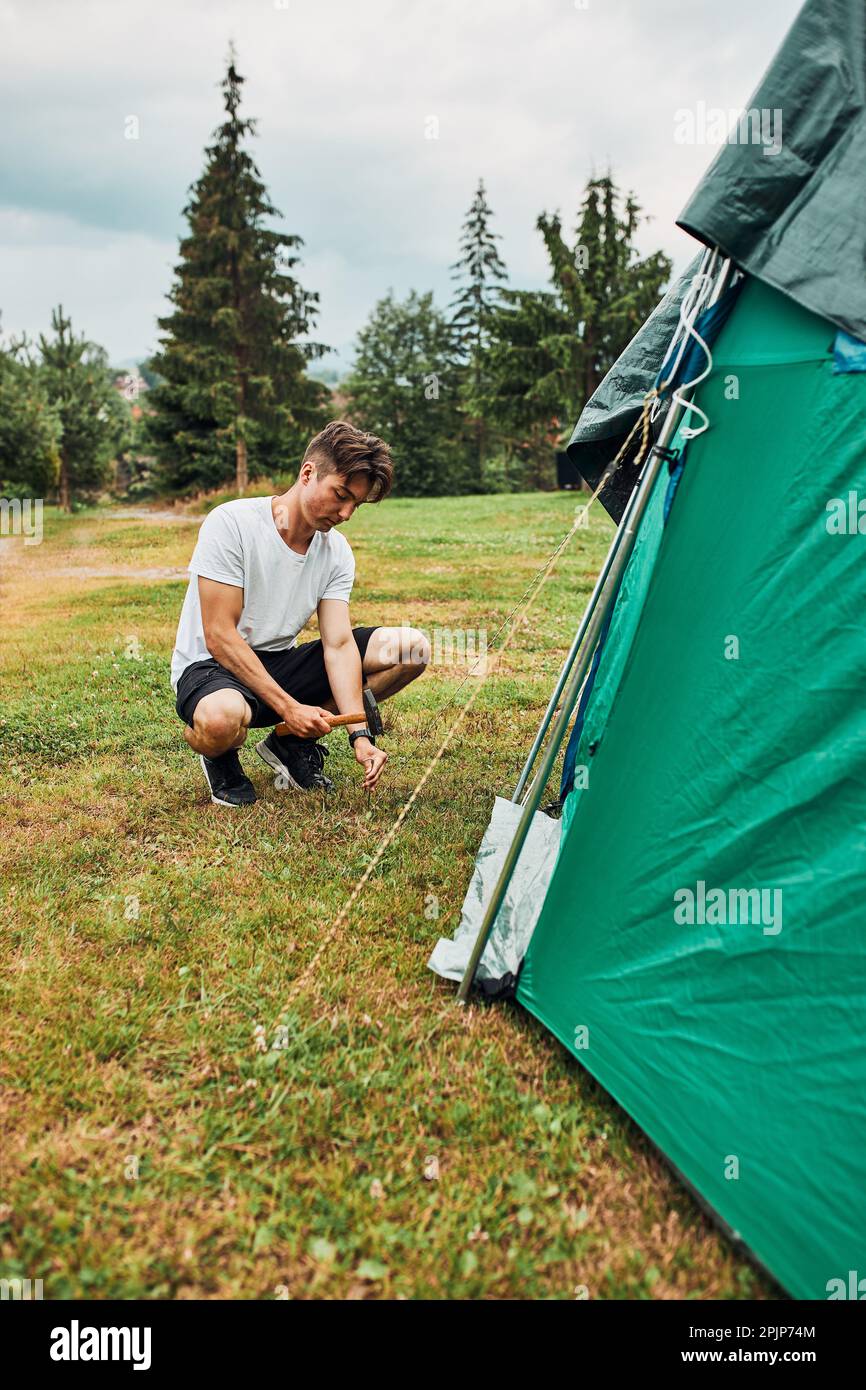 Young man putting up a tent on camping during summer vacation trip. Teenager putting the stakes into grassy ground using hammer Stock Photo