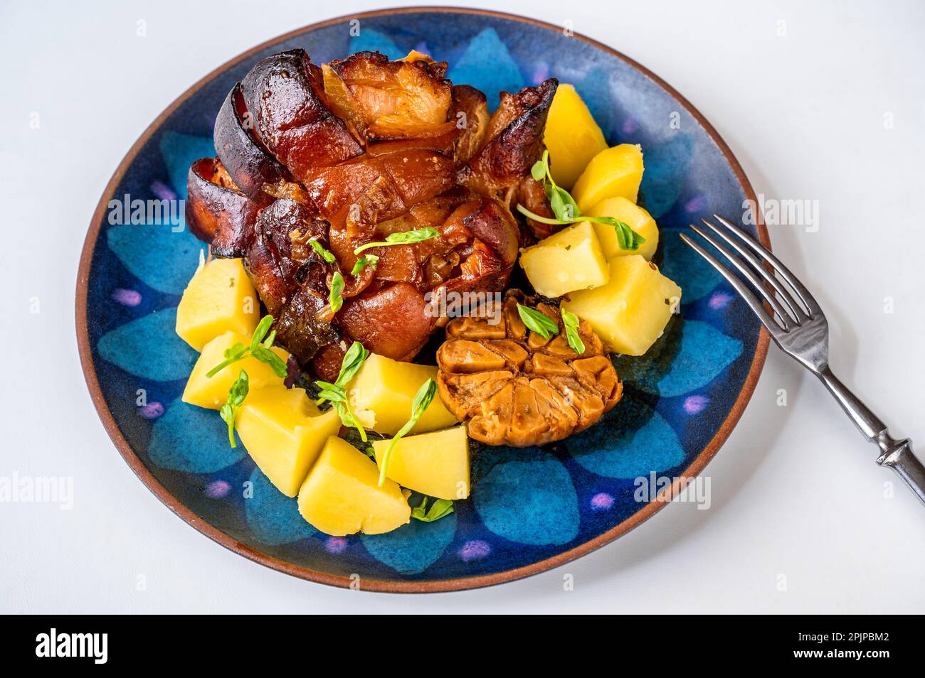 Baked until golden brown spicy pork belly, garlic and potato on decorative blue plate, fork on white background. Stock Photo
