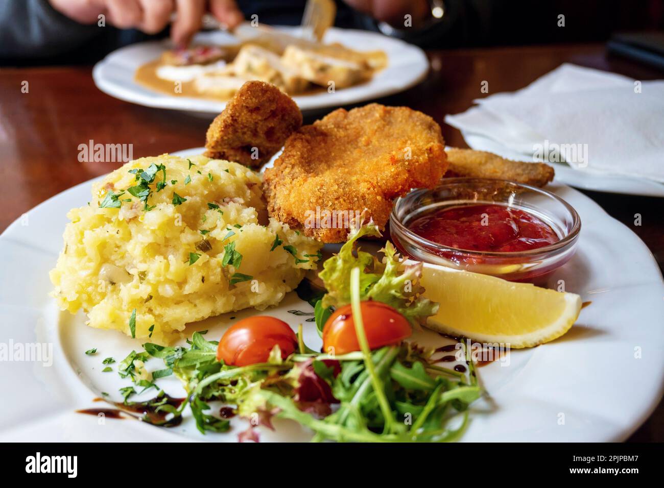 Fried schnitzel, mashed potato, green leaf, tomato and cranberry marmelade on white table, restaurant. Stock Photo