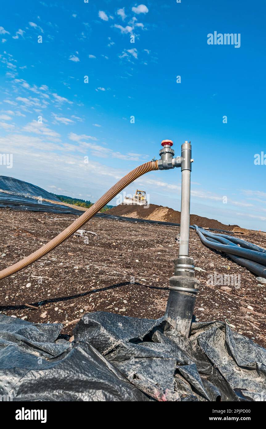 A vertical landfill methane gas wellhead and valve at an active landfill.  The hose and well pipe frame a soil compactor bulldozer in the background. Stock Photo
