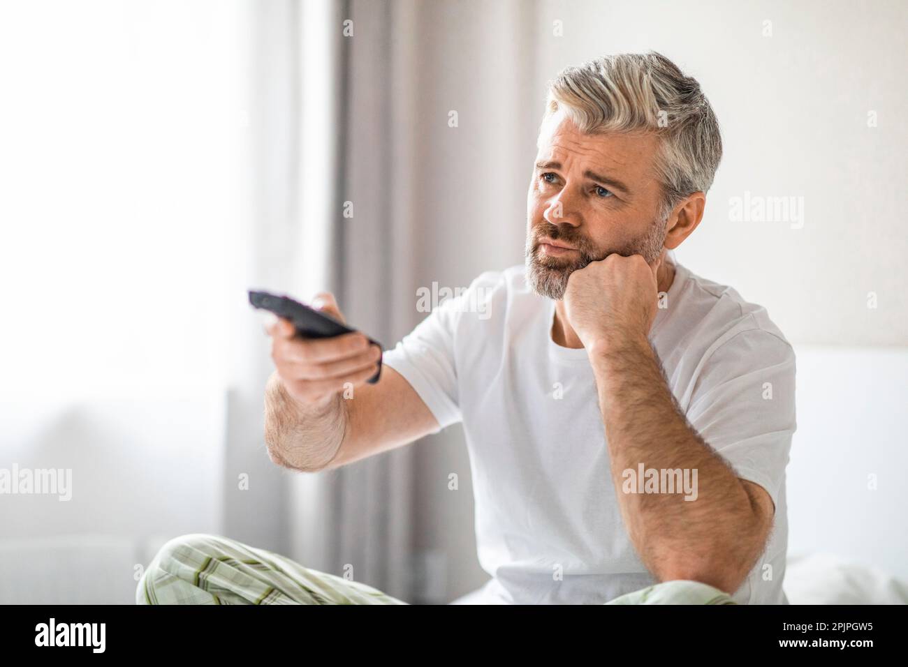 Bored mature man sitting on bed at home, watching tv Stock Photo