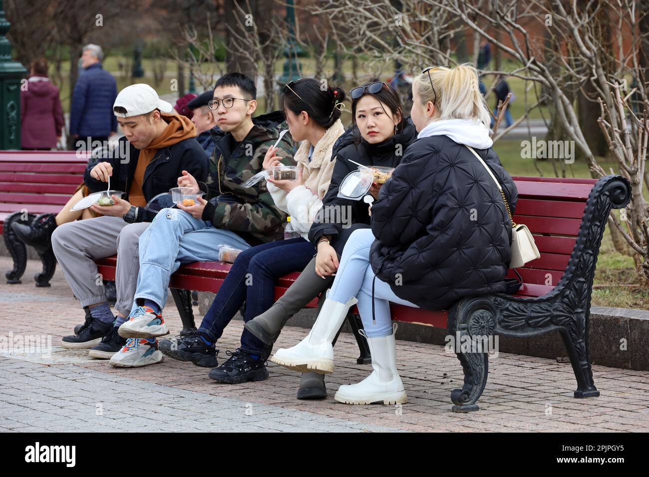 Asian girls and guys eat street food sitting on the bench in spring park. Tourists in Russia Stock Photo