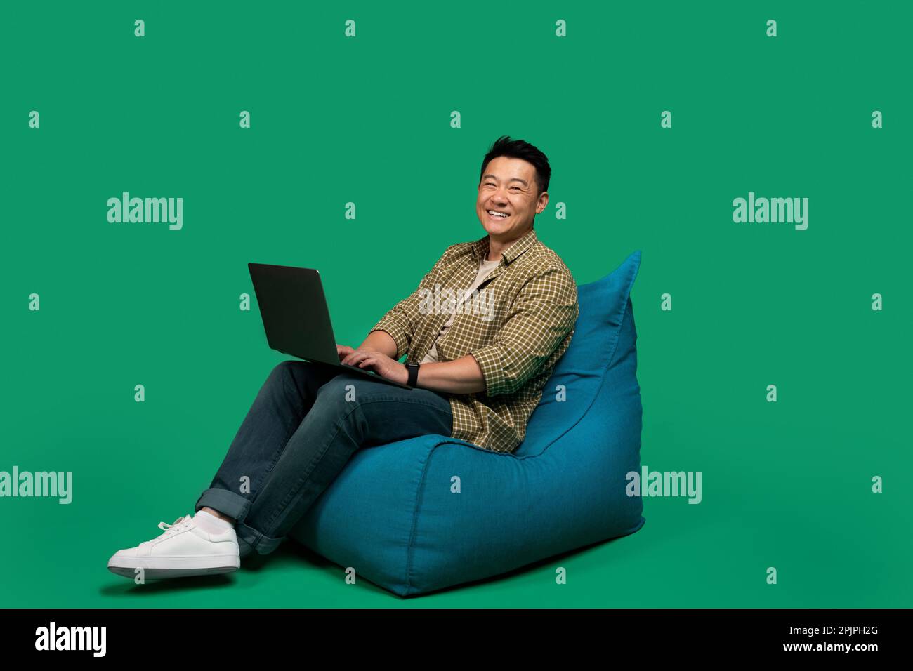 Happy asian middle aged man using laptop, working online while sitting in beanbag chair on green background, free space Stock Photo