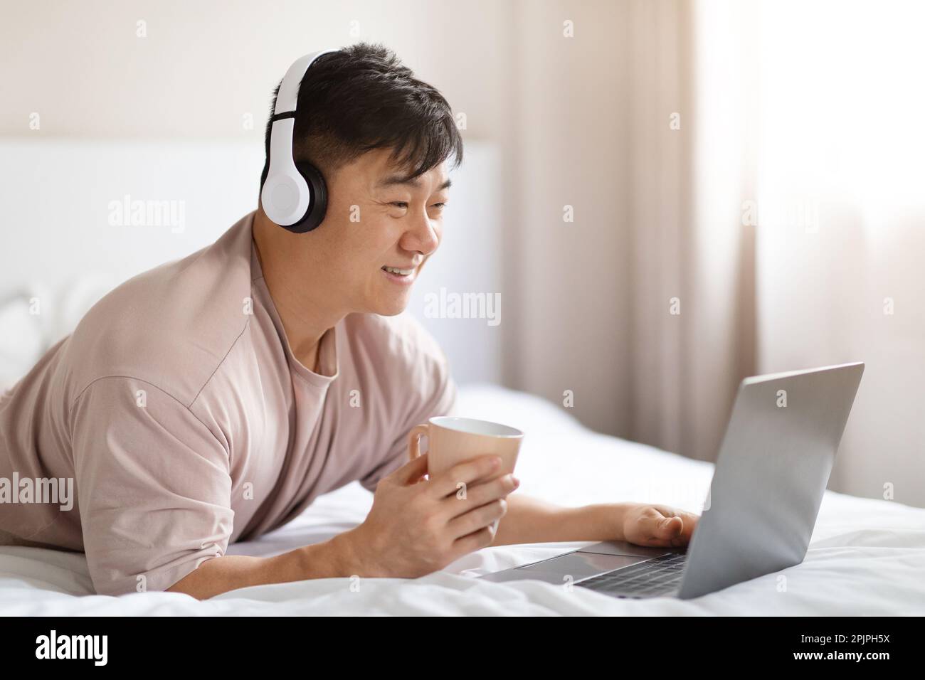 Positive asian man using laptop and headset in bed Stock Photo