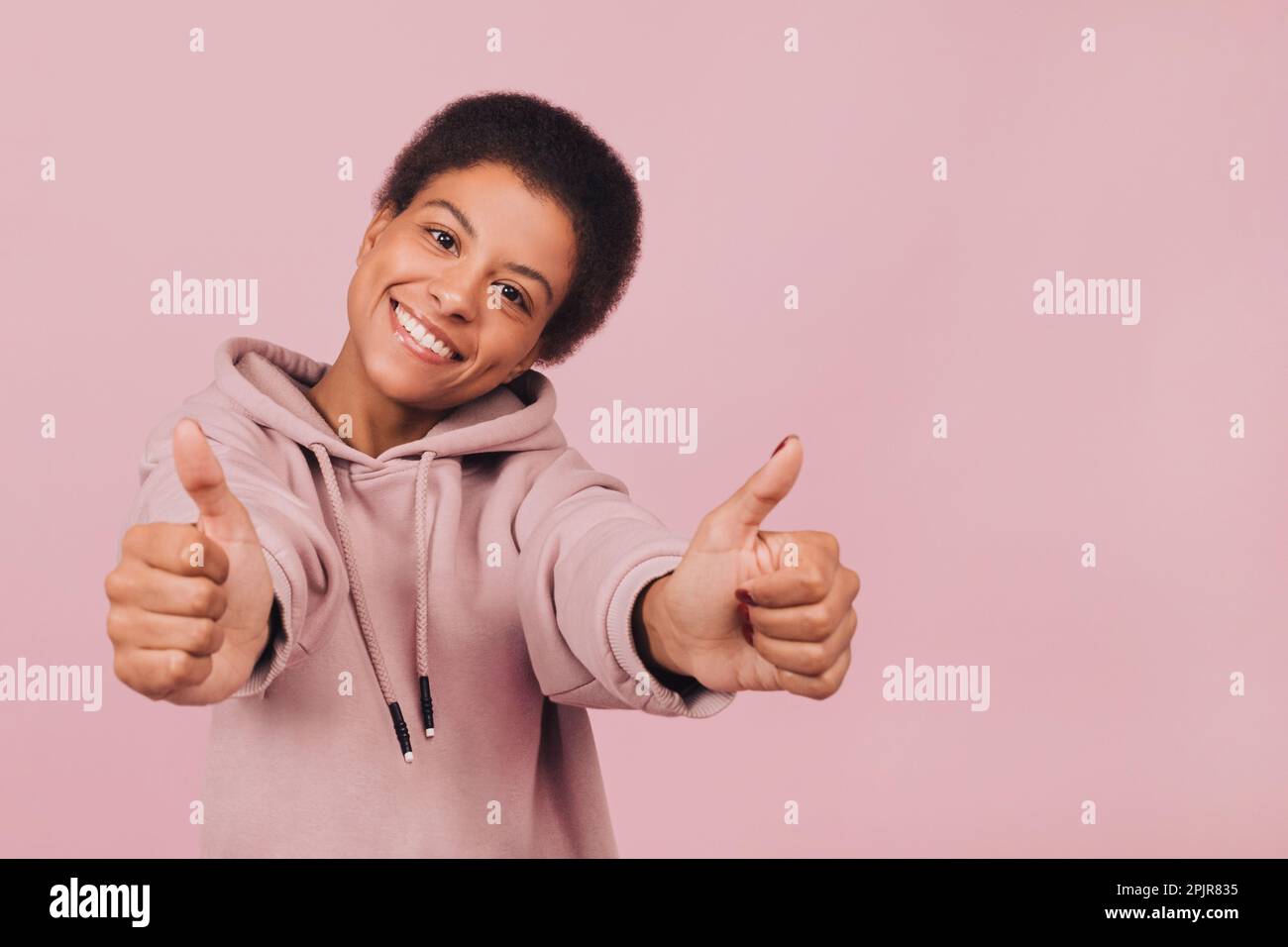 Close up portrait of happy young black woman showing thumbs up. Cheerful girl in casual clothes posing over pink background Stock Photo