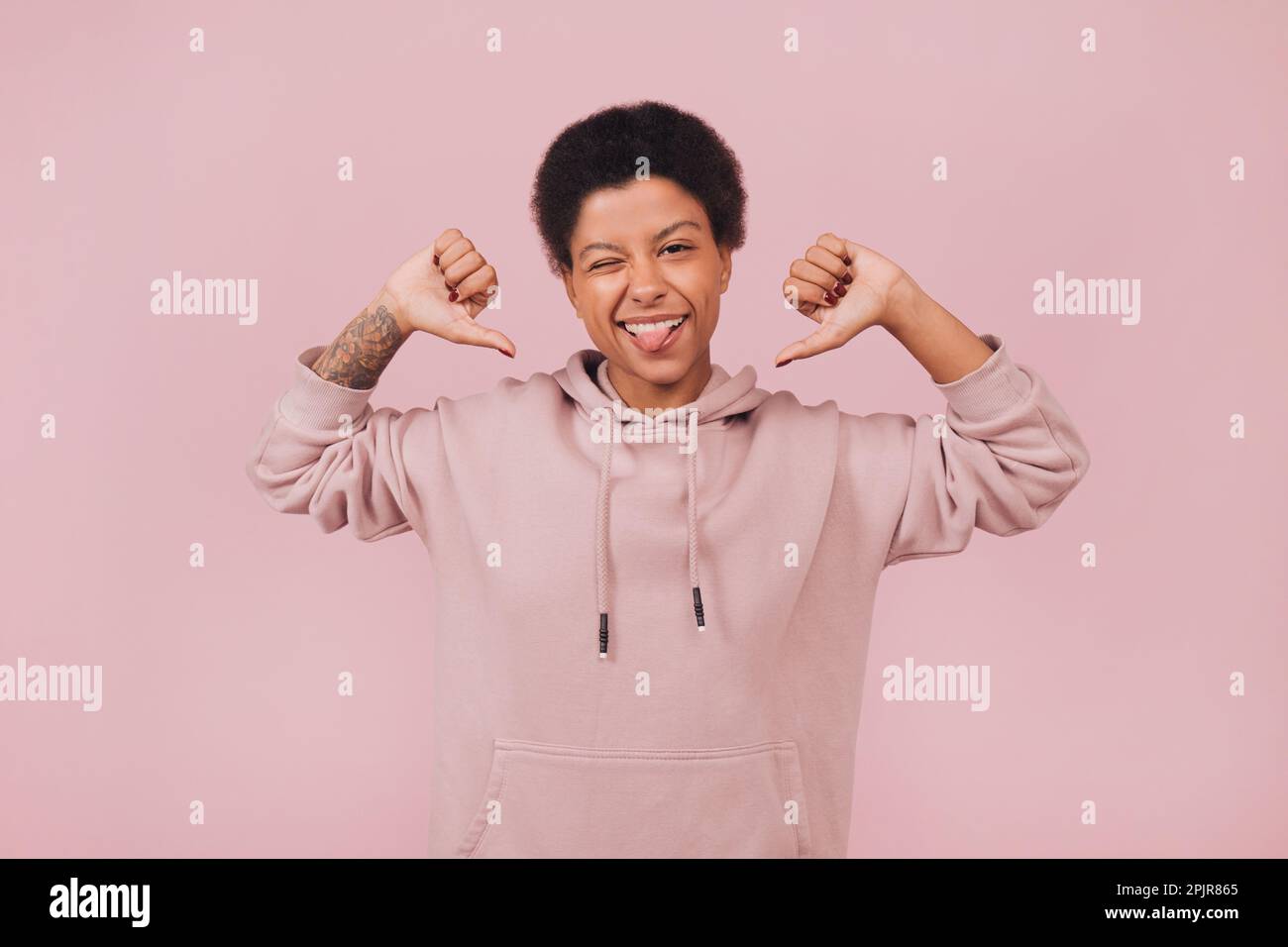 Joyful young black woman winking and showing on herself by thumbs. Cheerful student girl wearing hoodie posing on pink backdrop. Close up portrait Stock Photo