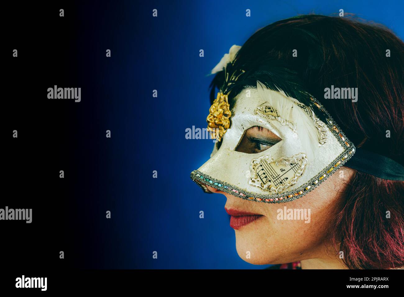 Portrait of a caucasian red head adult woman wearing a venetian theater mask, studio shoot blue baclground with selective focus Stock Photo