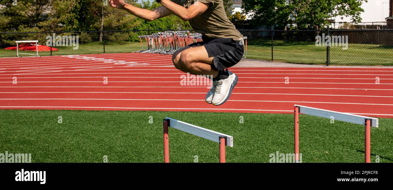 Seide view of a male athlete in shorts jumping over track hurdle on a turf field. Stock Photo