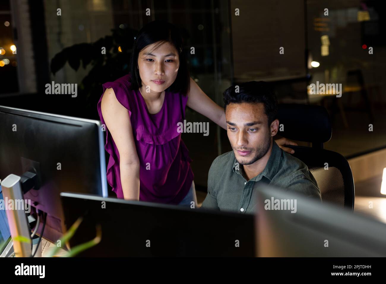 Diverse business people sitting at desk, using computer together, working late at office Stock Photo