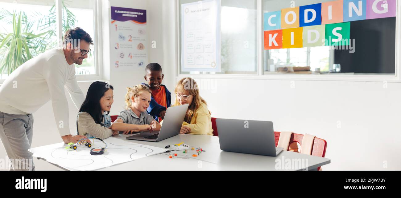 Primary school children learn to program robots with their teacher's guidance in a classroom. Group of kids using a laptop to develop a code that can Stock Photo