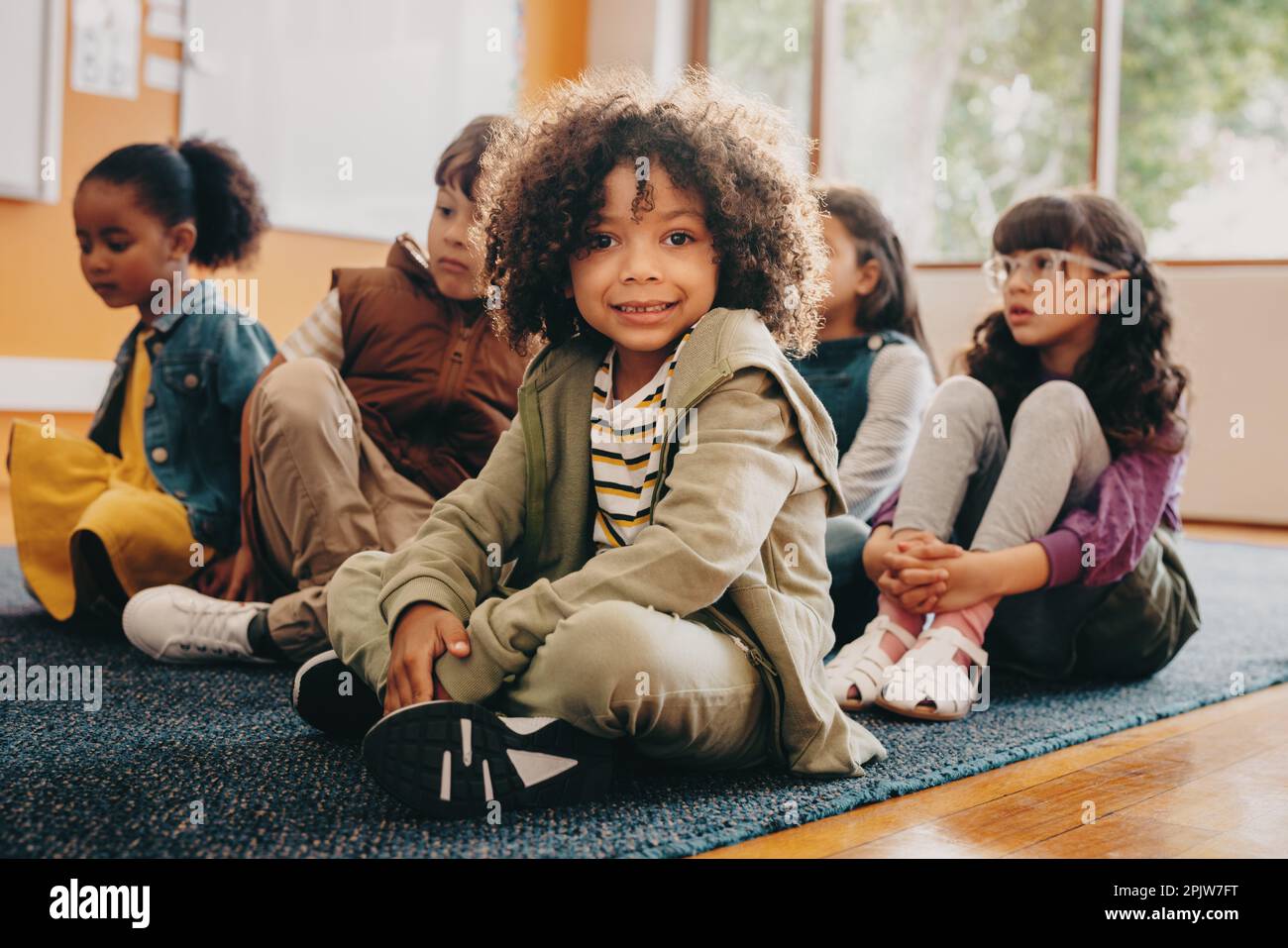 Boy sits in an elementary school class, he is looking at the camera. Multiracial kid attends a co-ed primary school with a group of multiethnic childr Stock Photo
