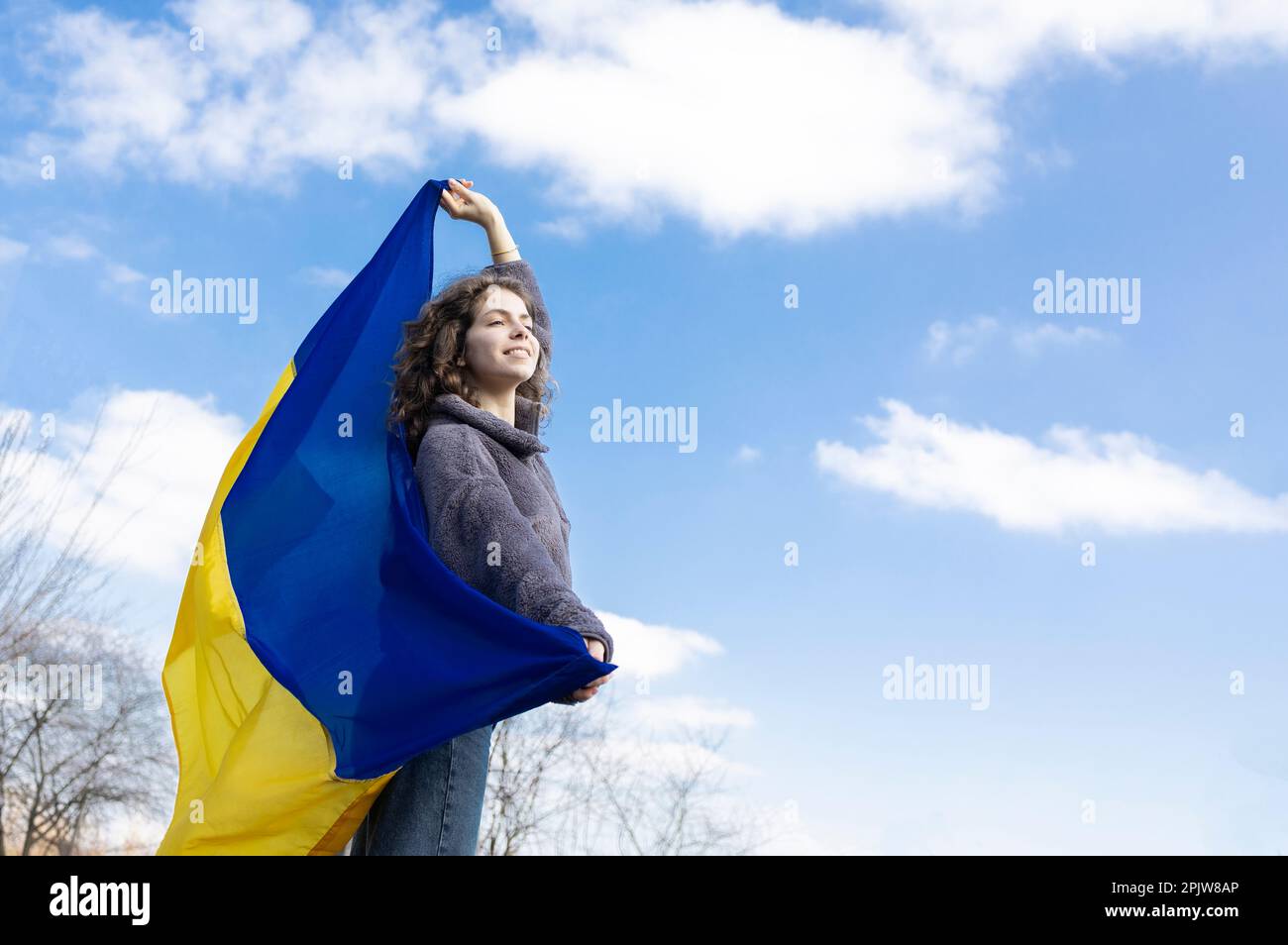 an adult girl of 18 years old, holds a Ukrainian yellow-blue flag behind her back against the sky, looks ahead, in the hope of a brighter future. Ukra Stock Photo