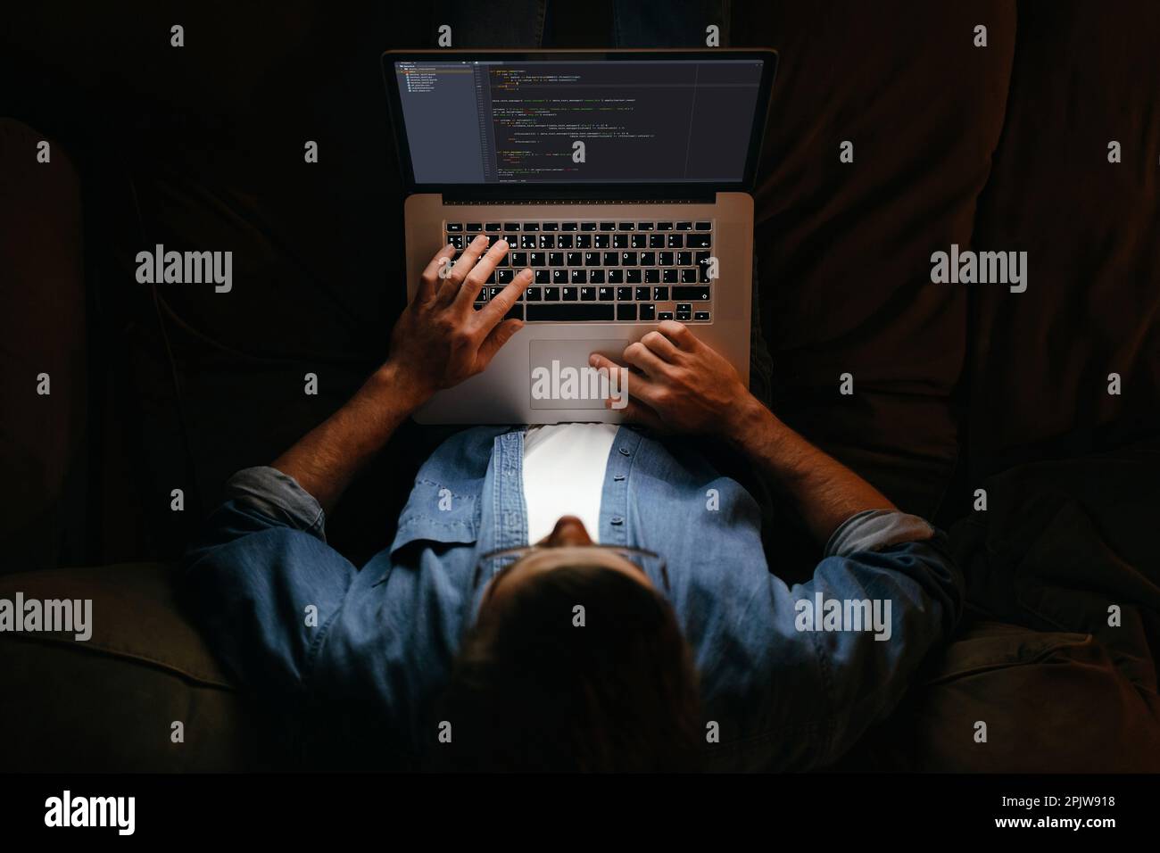 Young coder in eyeglasses is working remotely from home late at night, looking on display, typing code. Hard worker, programmer working overtime, taking work at home, has deadline.  Stock Photo