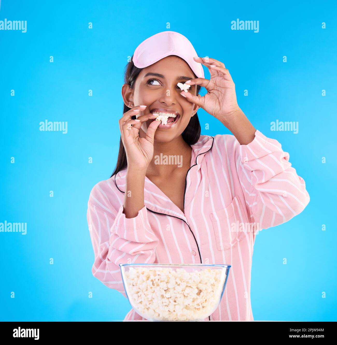Popcorn, pajamas and playful with a woman on a blue background in studio watching a movie for entertainment. Fun, video and night with an attractive Stock Photo