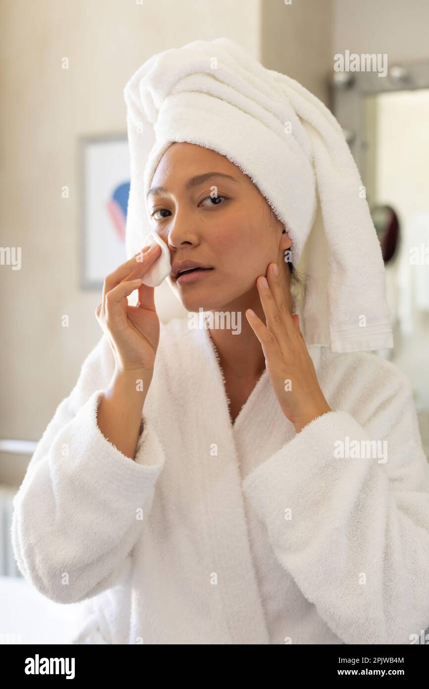 Happy asian woman wearing towel on head looking in bathroom mirror cleansing face using cotton
