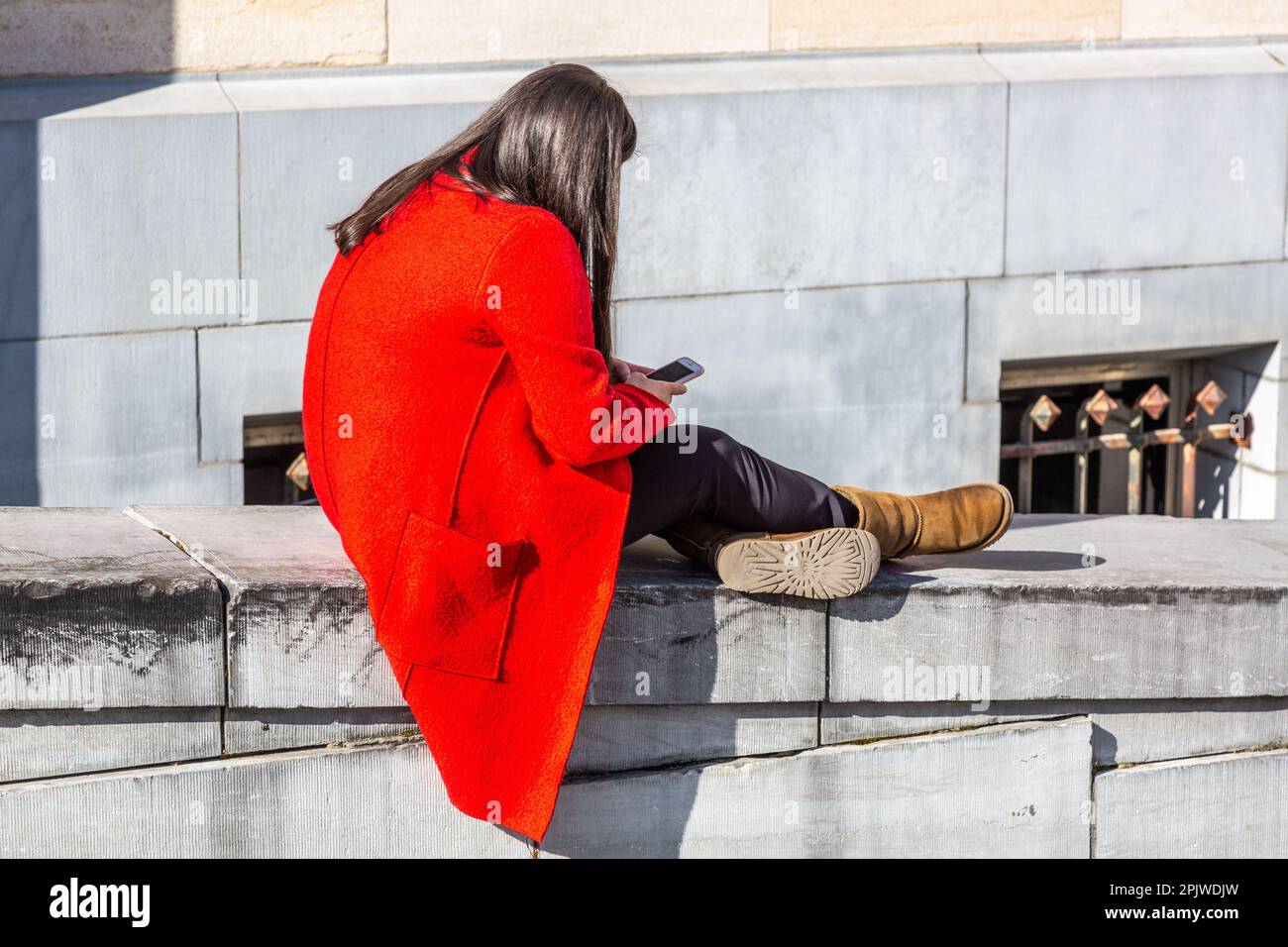 Young woman in a bright red coat looking at her phone, sitting on a parapet. Mont des Arts, Brussels. Stock Photo